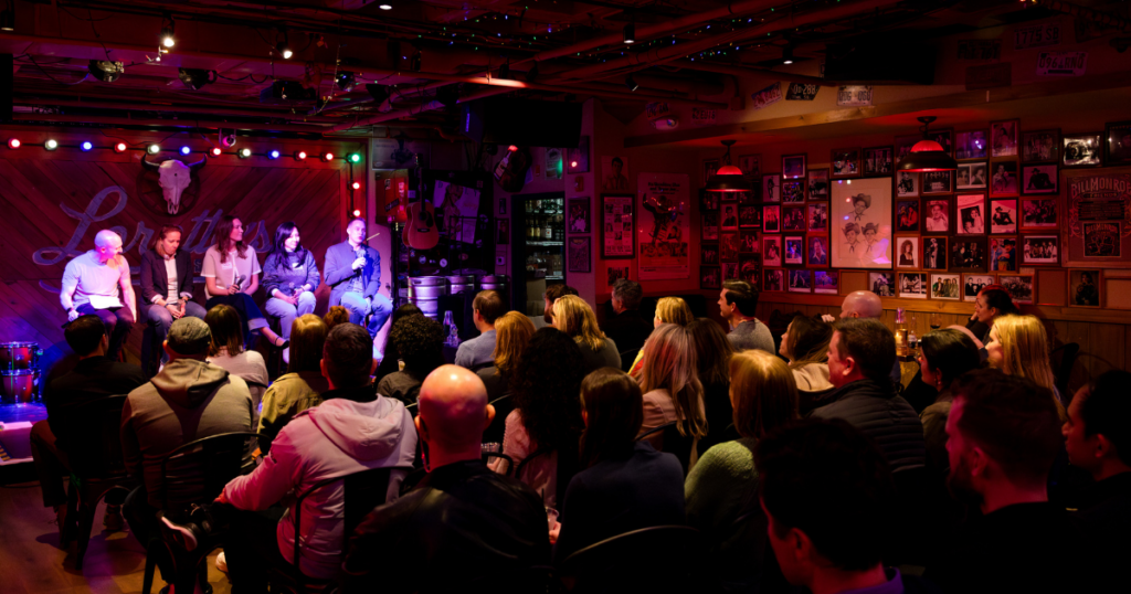 Wide-angle view of a B2B marketing expert panel discussion on stage at Loretta's bar venue during an Exit Five community meetup, full seated audience visible on both sides