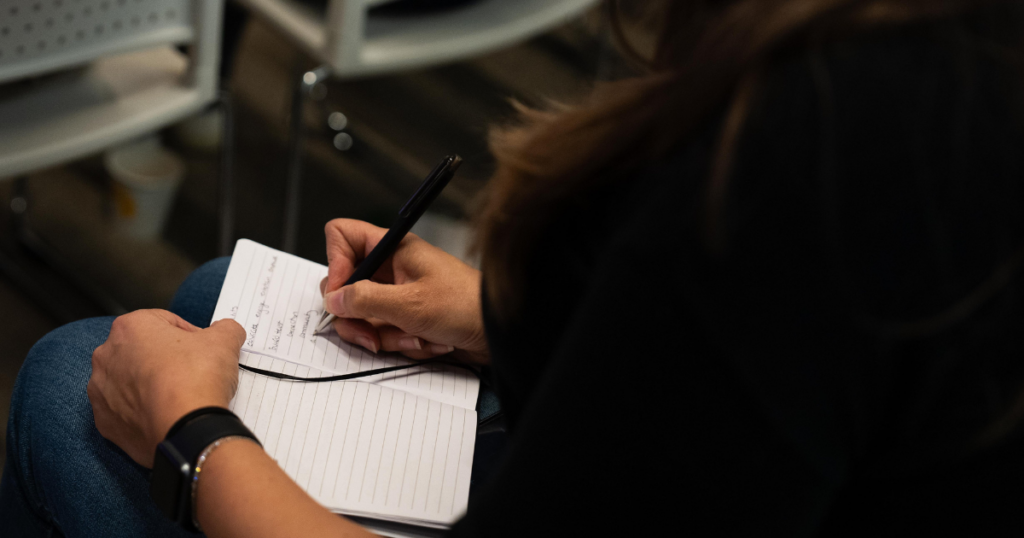 Close-up of a B2B marketer's hands writing notes in a lined notebook during a marketing conference session, smartwatch visible on wrist