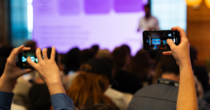 Audience members at a B2B marketing event raising smartphones to record a speaker's presentation projected on a purple-lit screen