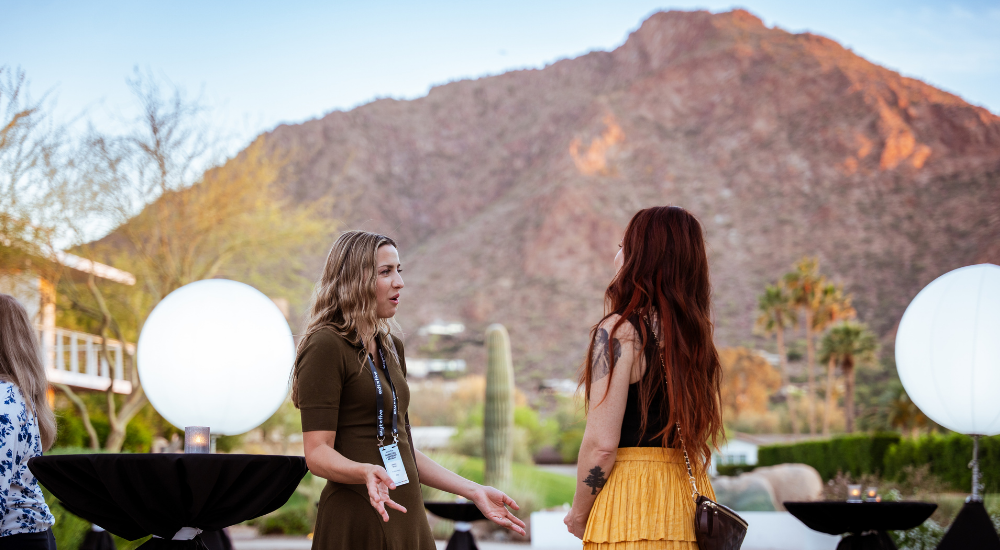 Two women talking outdoors, mountain backdrop.