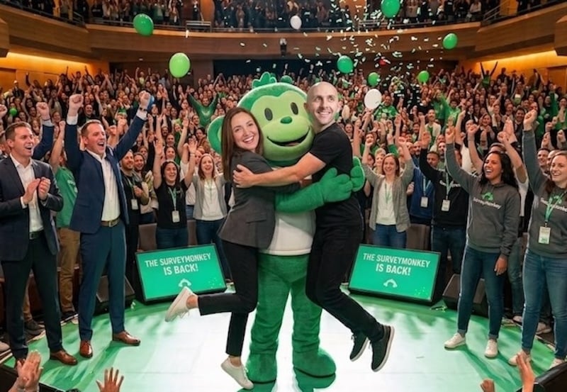 Town hall reenactment with two people hugging the green SurveyMonkey mascot as a crowd cheers and signs read The SurveyMonkey is back